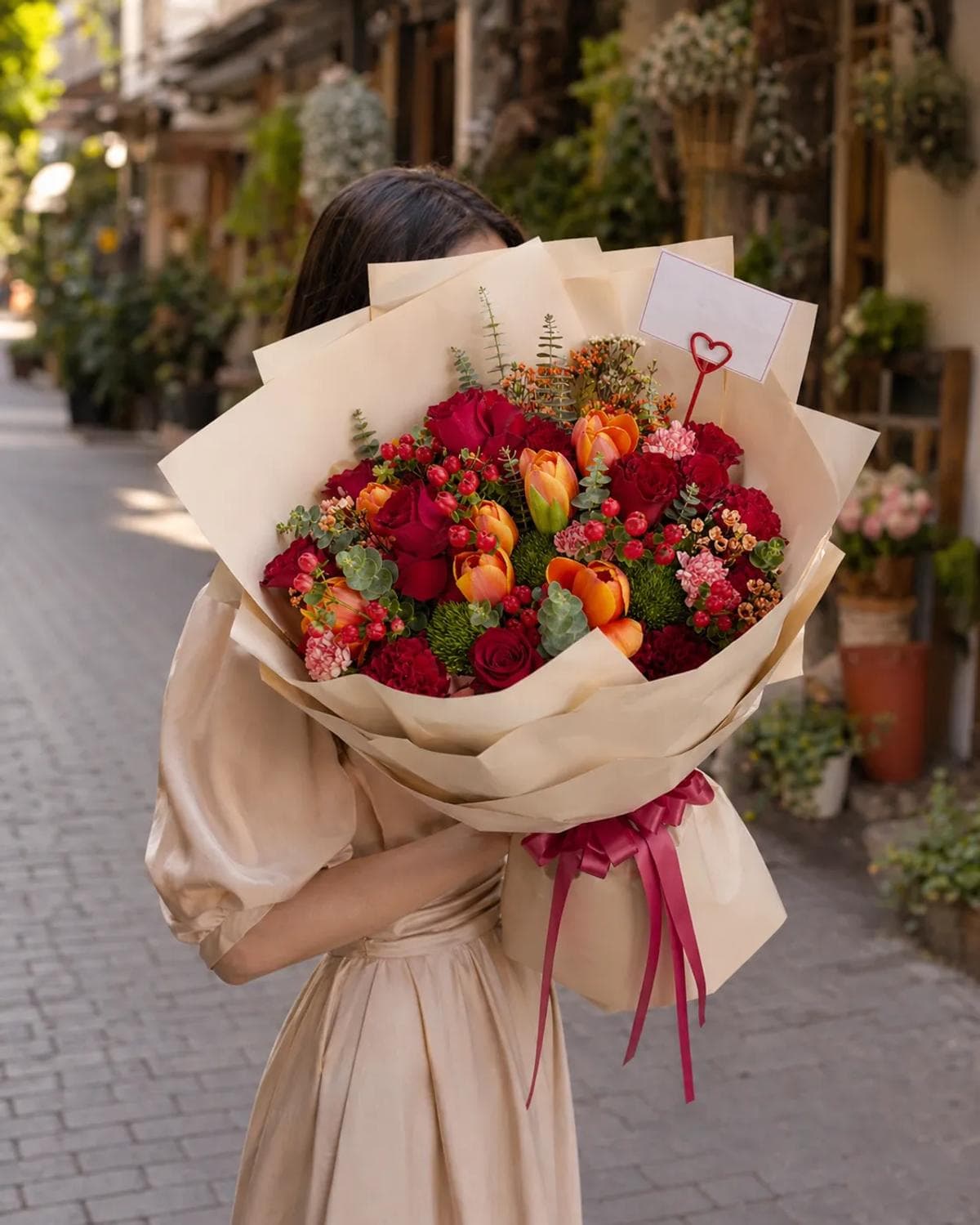 Celebrate life’s most vibrant moments with this stunning hand-tied bouquet. Featuring a sophisticated blend of deep red roses and sun-kissed orange tulips, this arrangement is accented with fresh berries and lush greenery for a textured, garden-fresh feel. Wrapped in elegant cream paper and finished with a classic burgundy ribbon, it is the perfect choice for anniversaries, birthdays, or any occasion that calls for a touch of timeless luxury and warmth.