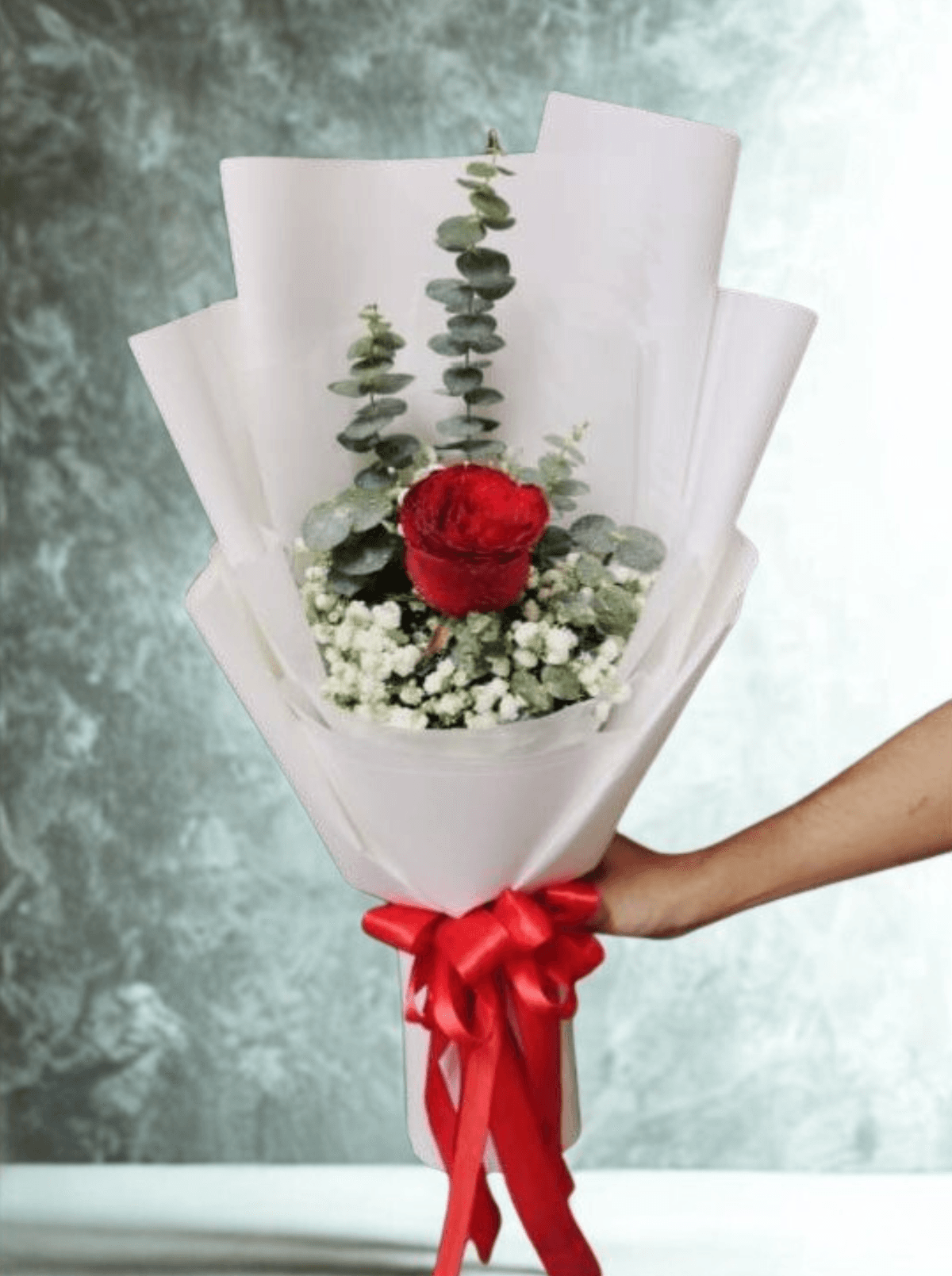 A minimalist single-rose bouquet with a bold romantic feel. One deep red rose stands at the center, softened by delicate white baby’s breath and fresh eucalyptus. Wrapped in clean white paper and finished with a bright red ribbon — simple, elegant, and perfect for a meaningful gesture.